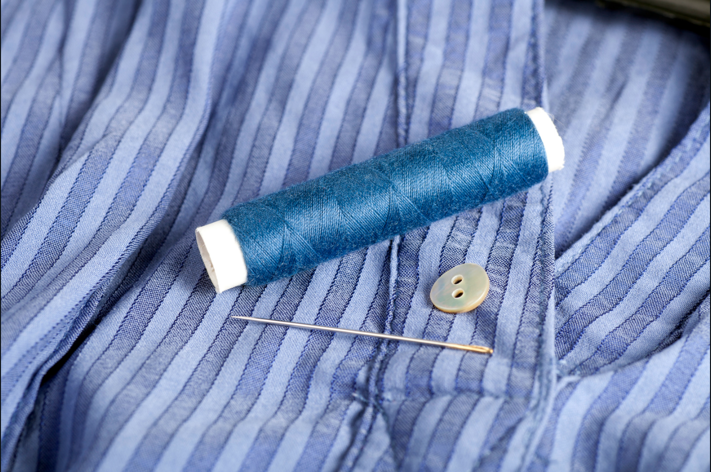 Blue pinstriped shirt and a spool of blue thread overtop. Next to that are a need and button indicating the button needs to be reattached to the shirt.