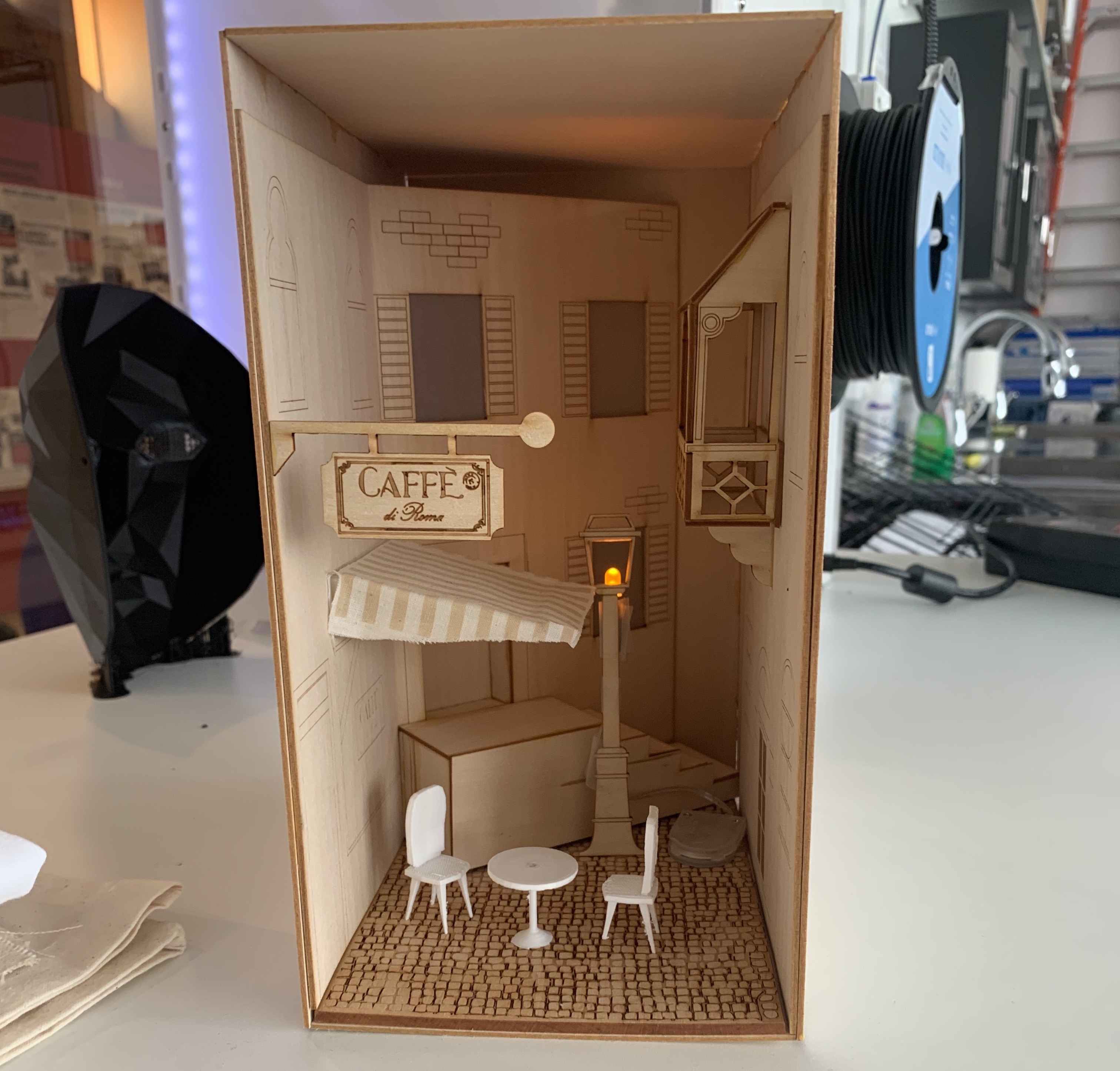 Wooden lasercut replica of an alley in a Eurpoean style. Within the replica is a sign that says cafe, 3D printed mini chairs and table, and windows.